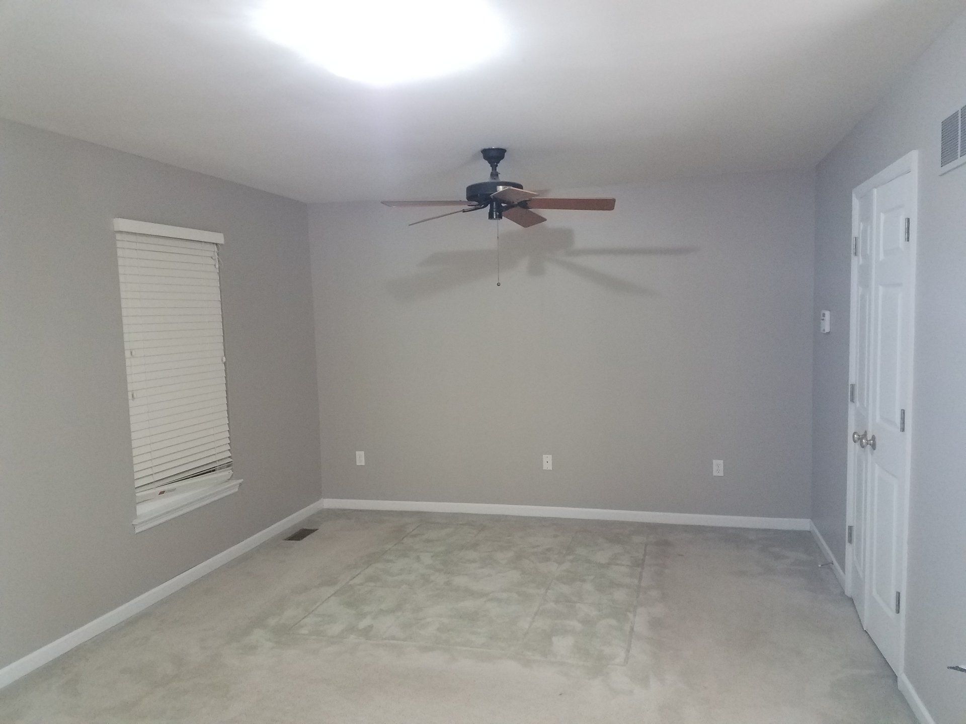 Empty room with gray walls, beige carpet, window with blinds, and ceiling fan. White door.