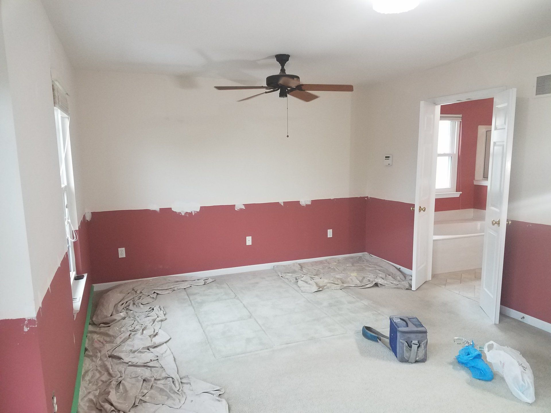 Room with burgundy lower walls, white upper walls. Ceiling fan, open doorway, and flooring covered in protective material.