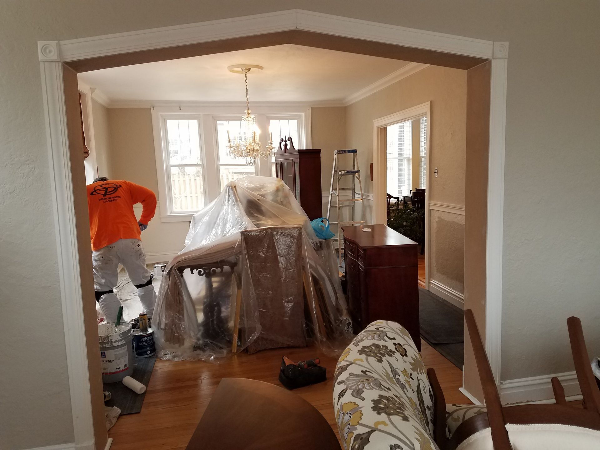 Interior room during painting, featuring a painter in orange, furniture covered in plastic, and doorway.