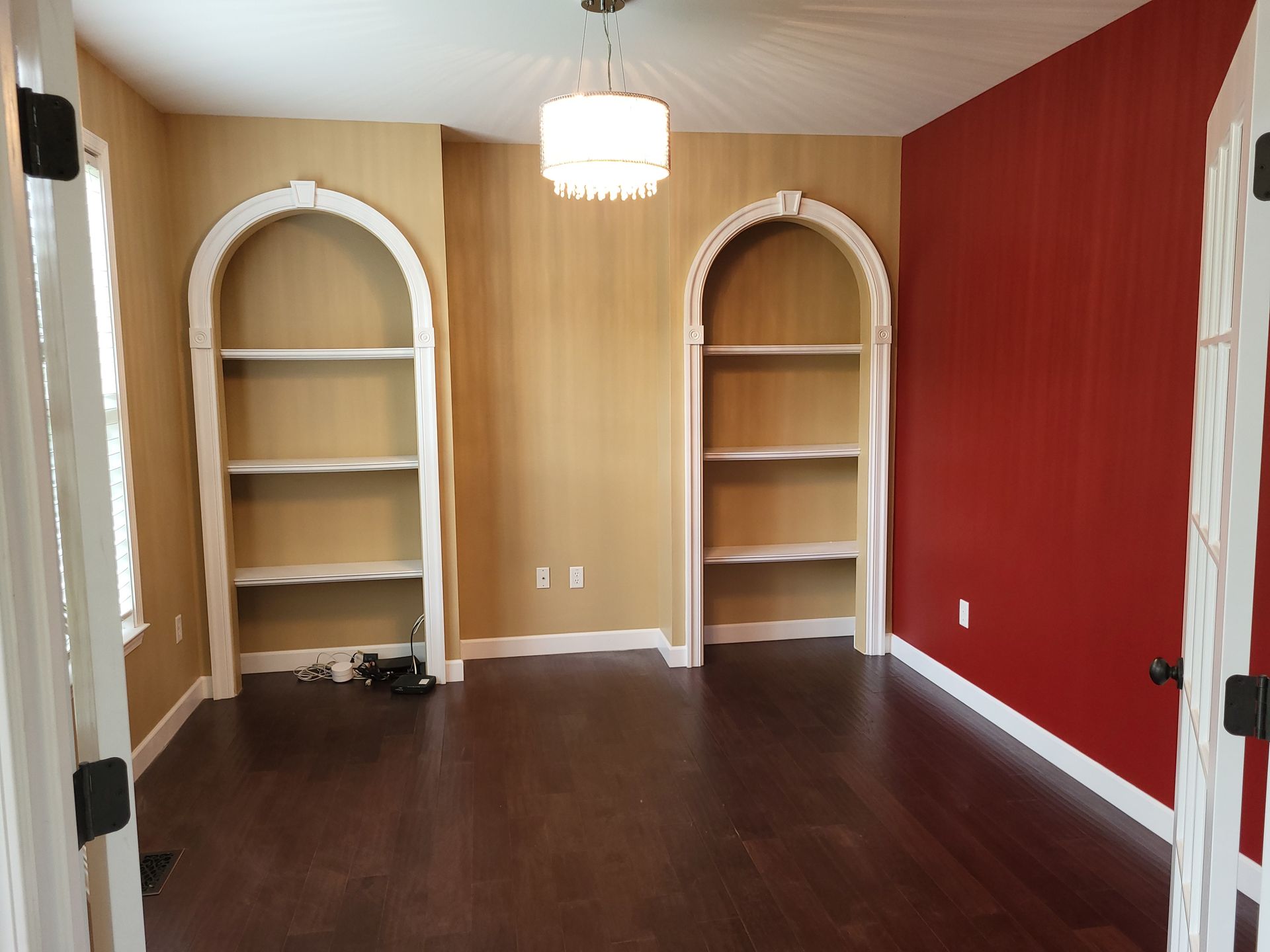 Room with dark wood floor, two arched bookshelves, and red/gold walls; chandelier.