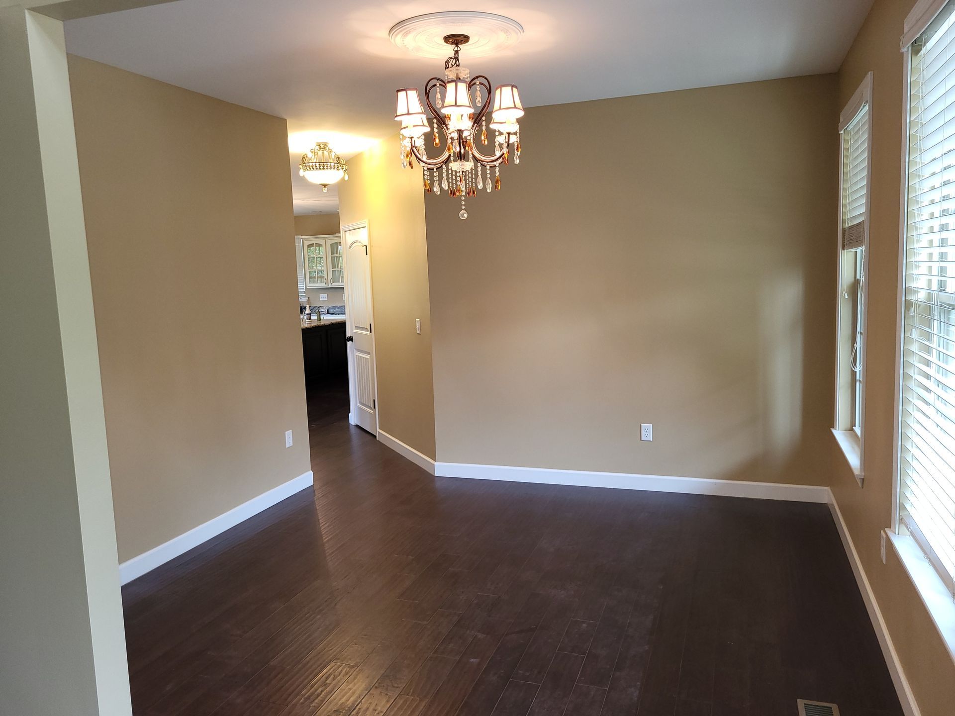 Empty dining room with dark wood floor, tan walls, chandelier, and a view of another room.