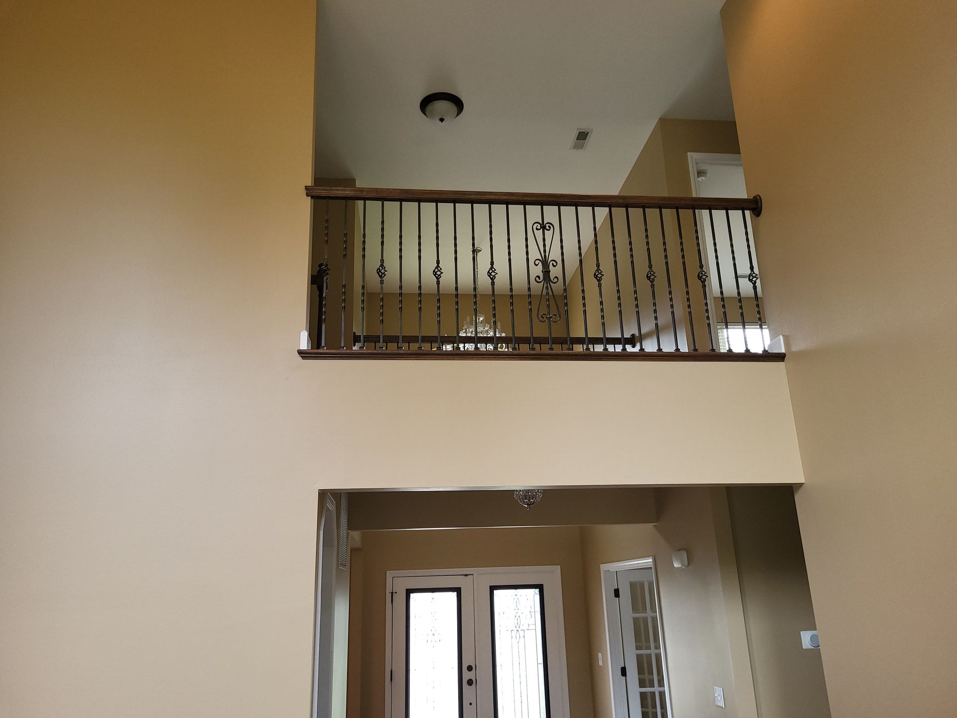 Interior view of a two-story foyer with a balcony and doorway; beige walls, dark railing.