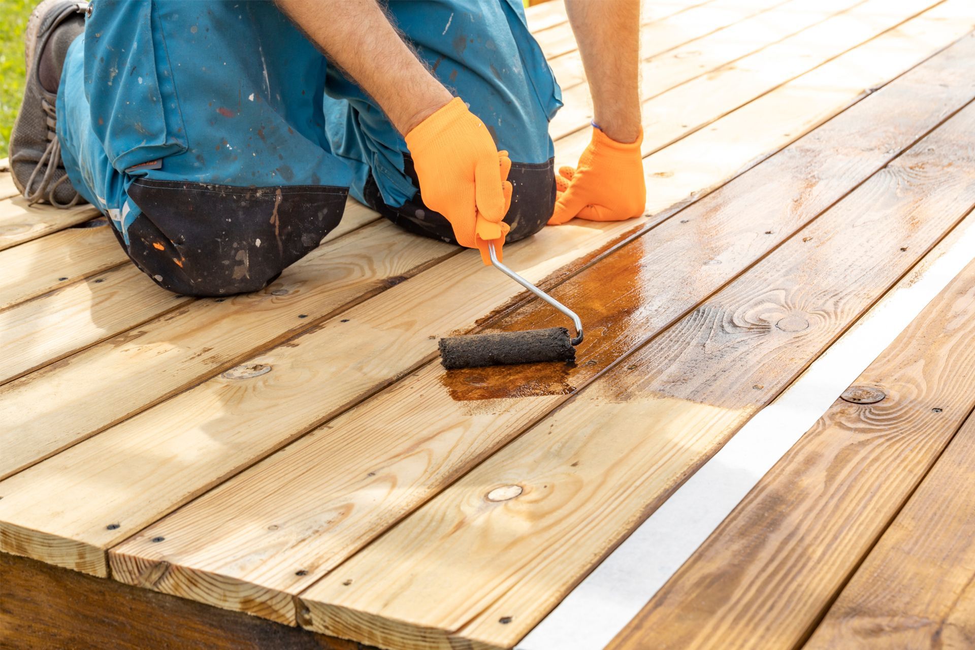 Person applying brown stain to a wooden deck with a paint roller, wearing orange gloves.