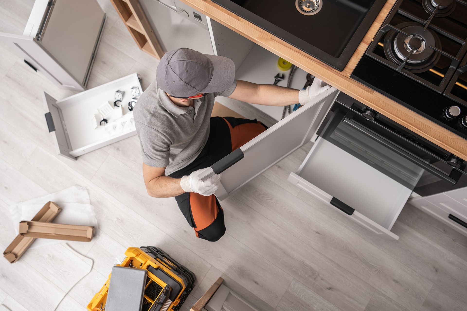Person kneeling, working under a kitchen cabinet, with open drawers and tools visible.