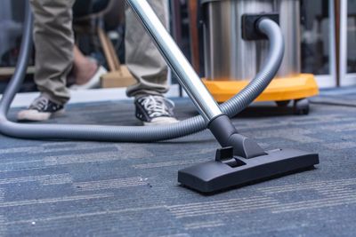 Person vacuuming a carpet with a gray hose and black floor nozzle outdoors