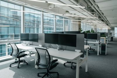 Modern open-plan office with white desks, black chairs, and large windows overlooking glass buildings