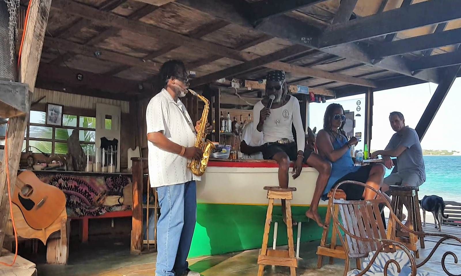 Saxophone player at a beach bar. People seated, listening. Blue water in the background.