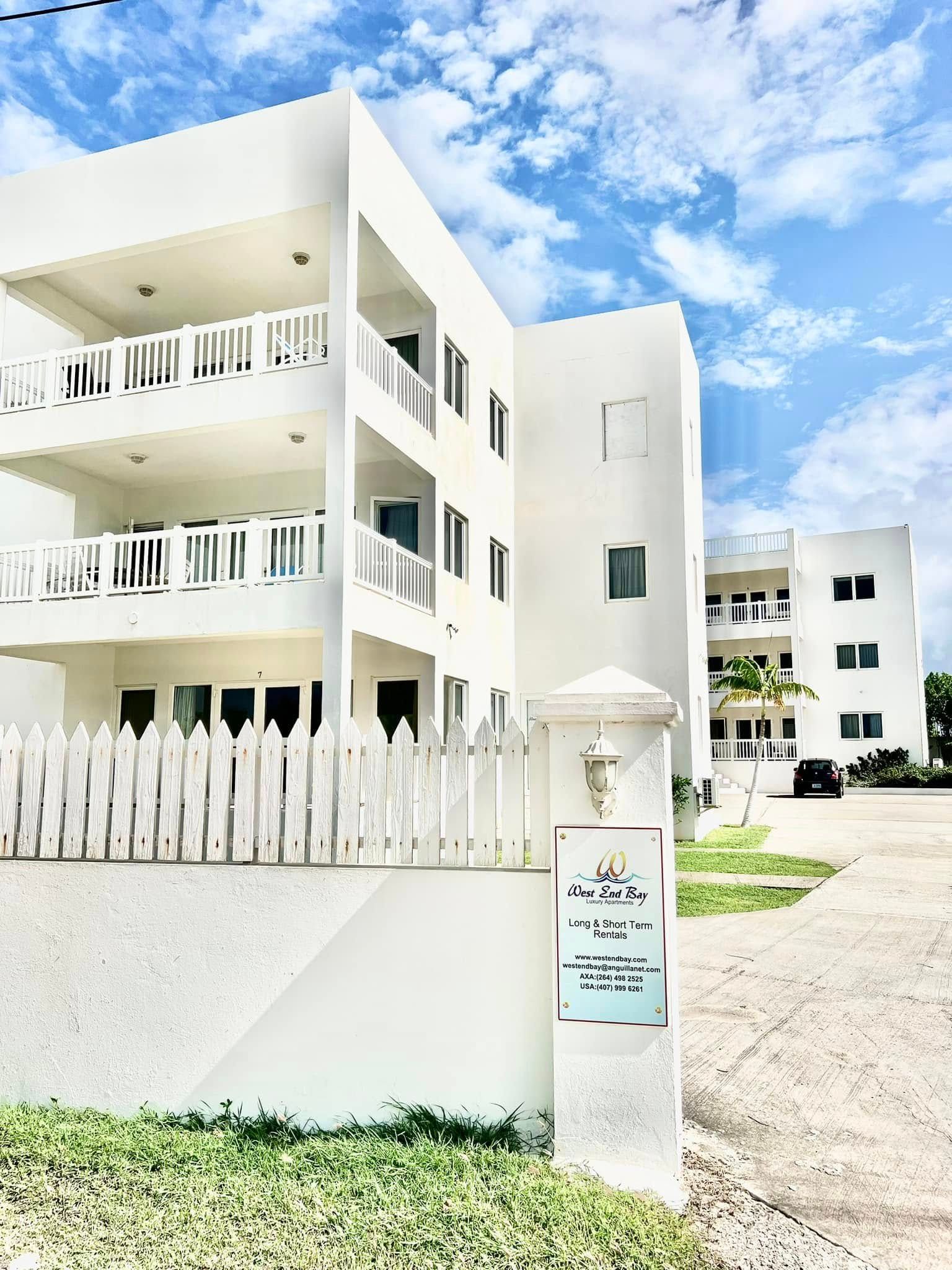 White multi-story apartment building with balconies, behind a white picket fence, under a blue sky.