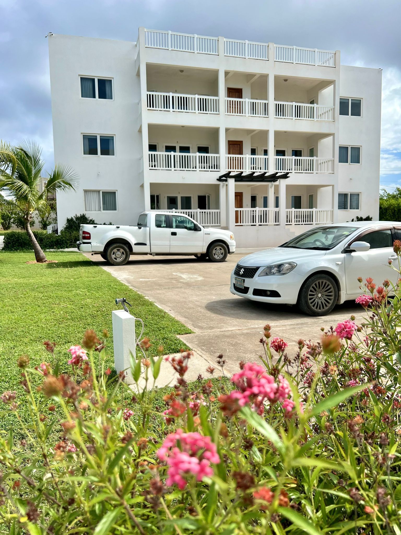 White apartment building with balconies, cars parked in front, and flowers in foreground.