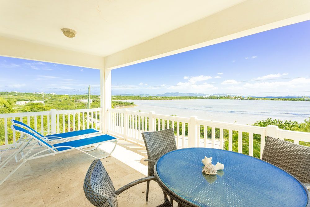 Balcony overlooking water, featuring blue chairs and table, white railing, and bright blue sky.