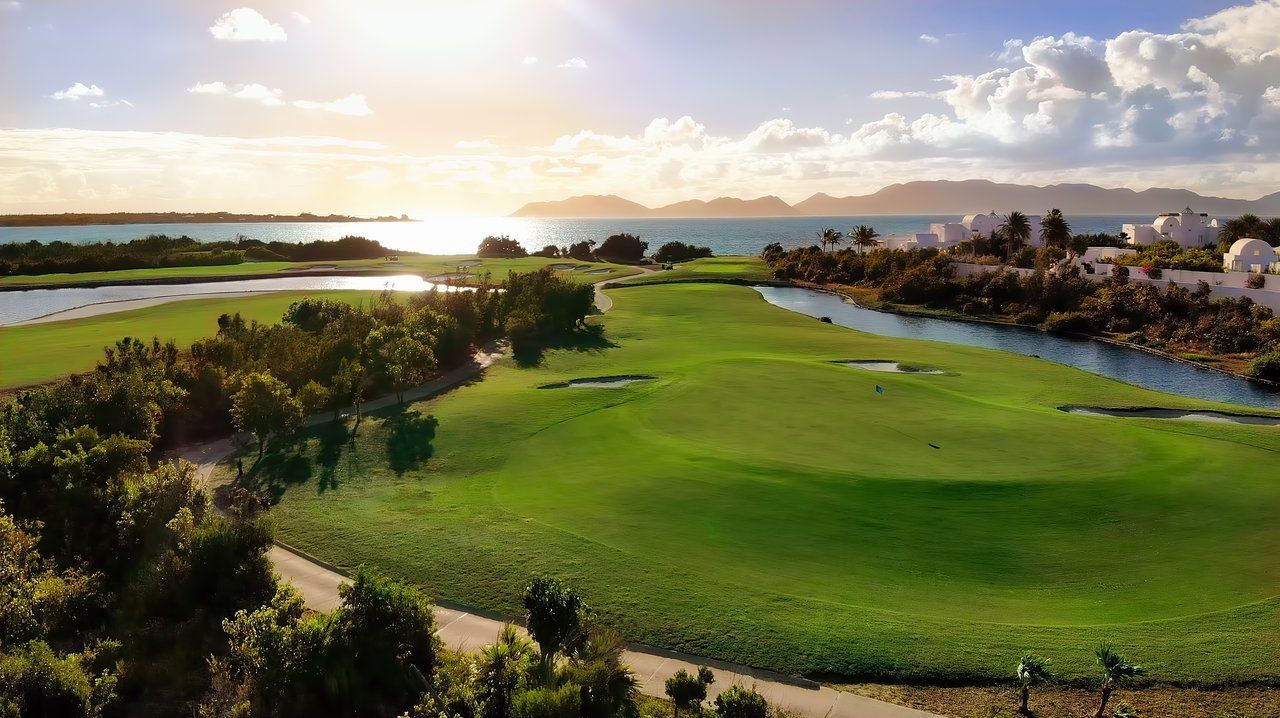 Green golf course beside water with buildings, under a sunny sky.