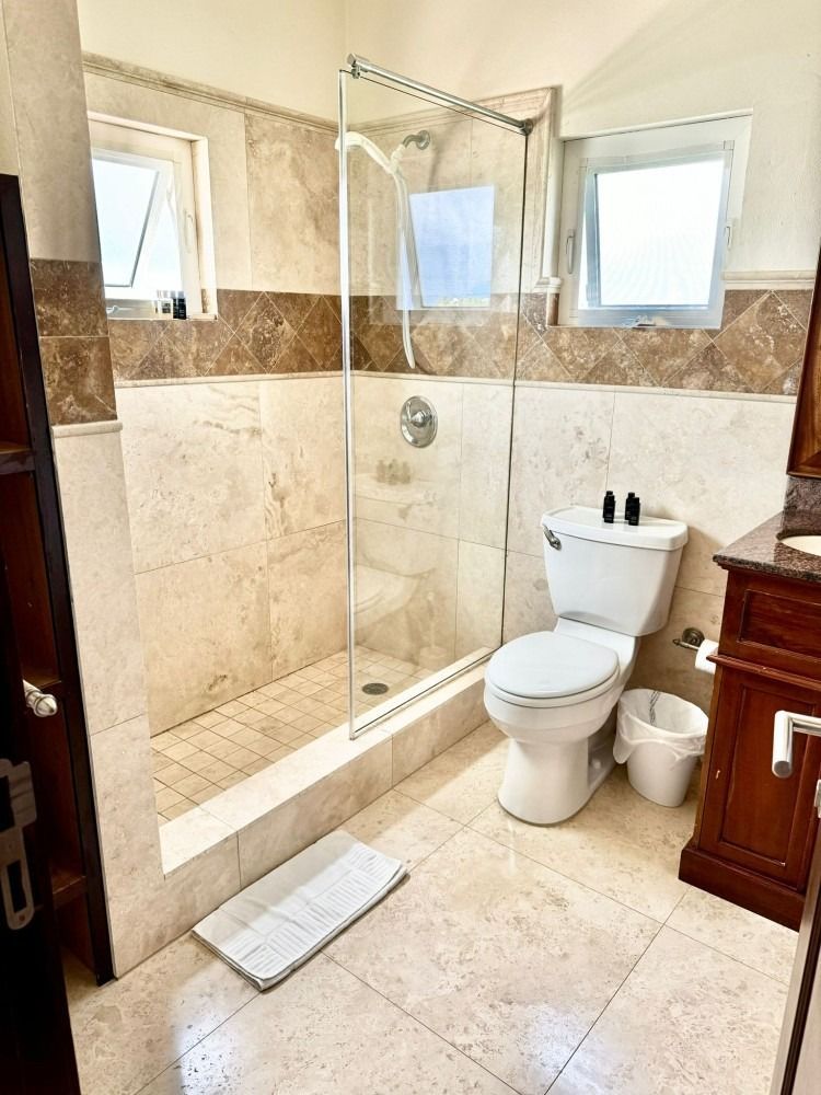 Bathroom with tiled walls and floor, glass shower, toilet, cabinet, and windows.