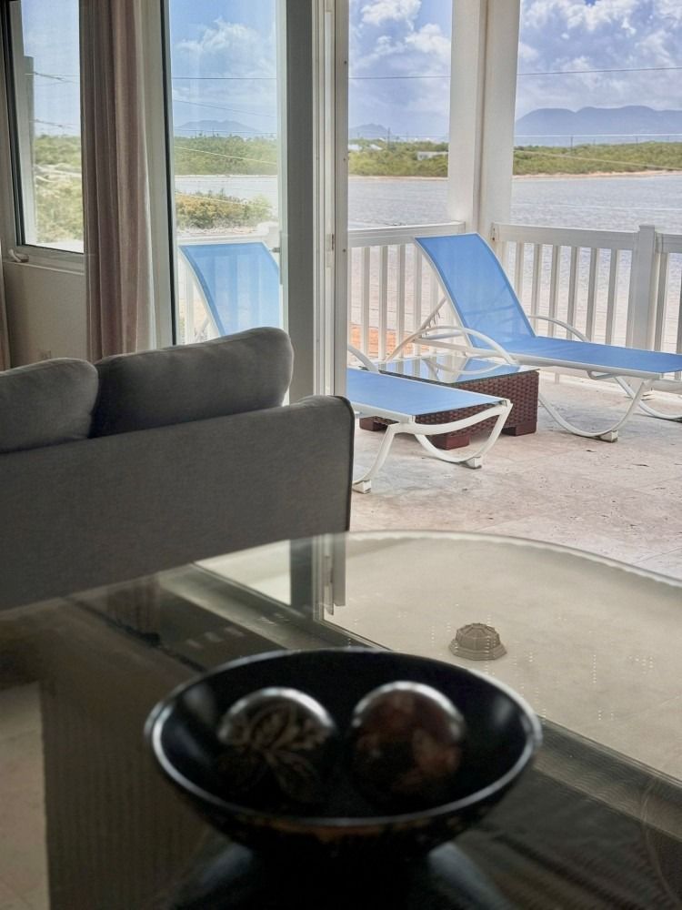 Living room with water view; grey couch, glass table, black bowl with spheres, blue lounge chairs on patio.