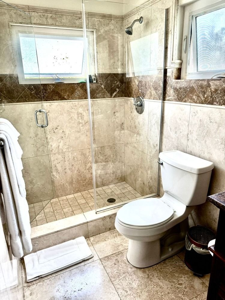 Bathroom with glass shower, toilet, and towel rack, featuring beige tile and a small window.