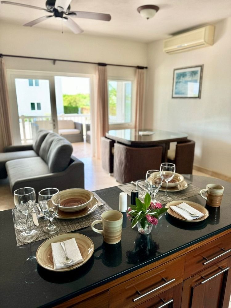 Dining area with table set, leading to a balcony and living area with gray sofa and kitchen island.