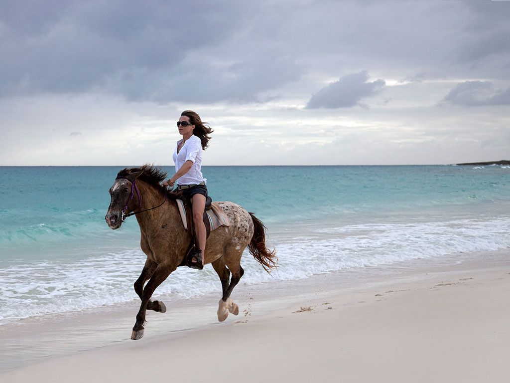 Woman riding a horse along a white sand beach, turquoise water, cloudy sky.