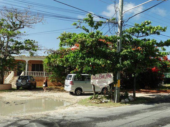A building and vehicles with a woman walking, partly obscured by greenery; a sign is present.