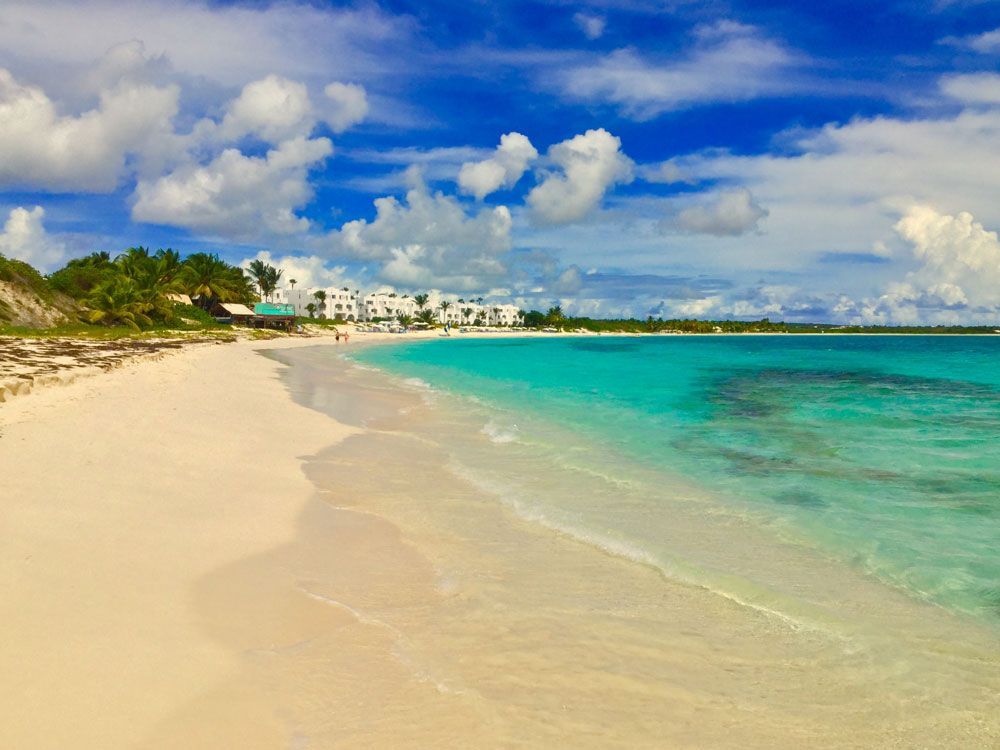White sand beach with turquoise water under a blue sky with fluffy clouds; white buildings line the shore.