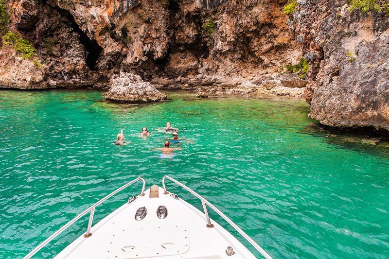 Boat in turquoise water near a rocky shore with people swimming.