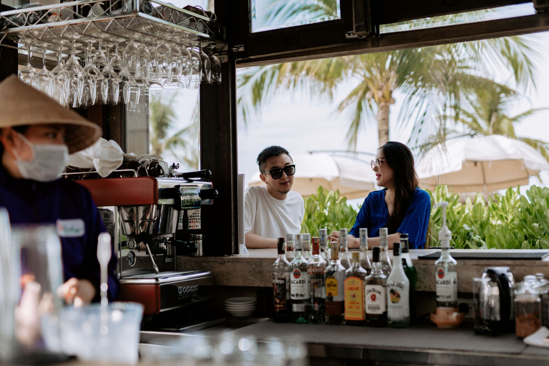 People talking at a beachside bar with bottles and hanging glasses in the foreground