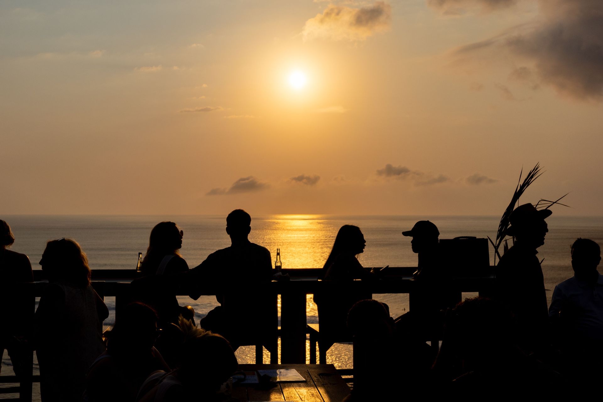 Silhouetted people seated by the beach at sunset, with the sun reflecting on the water.