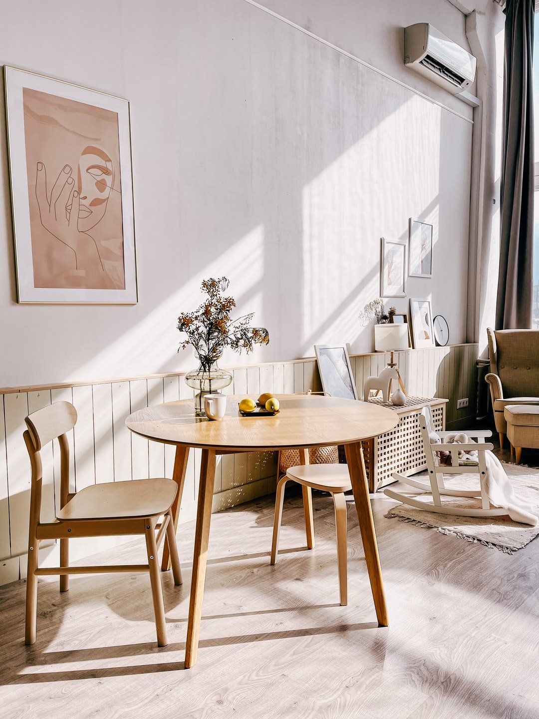 Sunlit dining nook with a round wooden table, chairs, flowers, and neutral decor by a bright window
