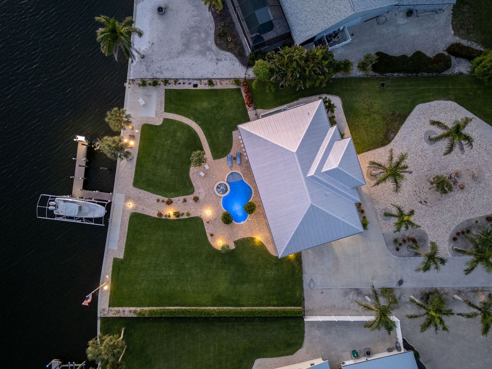 Aerial view of a waterfront home with a gray roof, blue pool, and palm trees beside a dock.