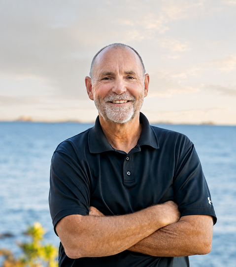 A man with a beard is standing with his arms crossed in front of the ocean.
