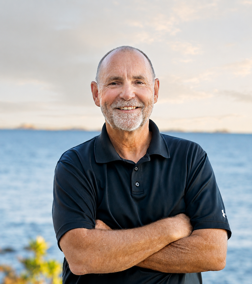 A man with a beard is standing with his arms crossed in front of the ocean.