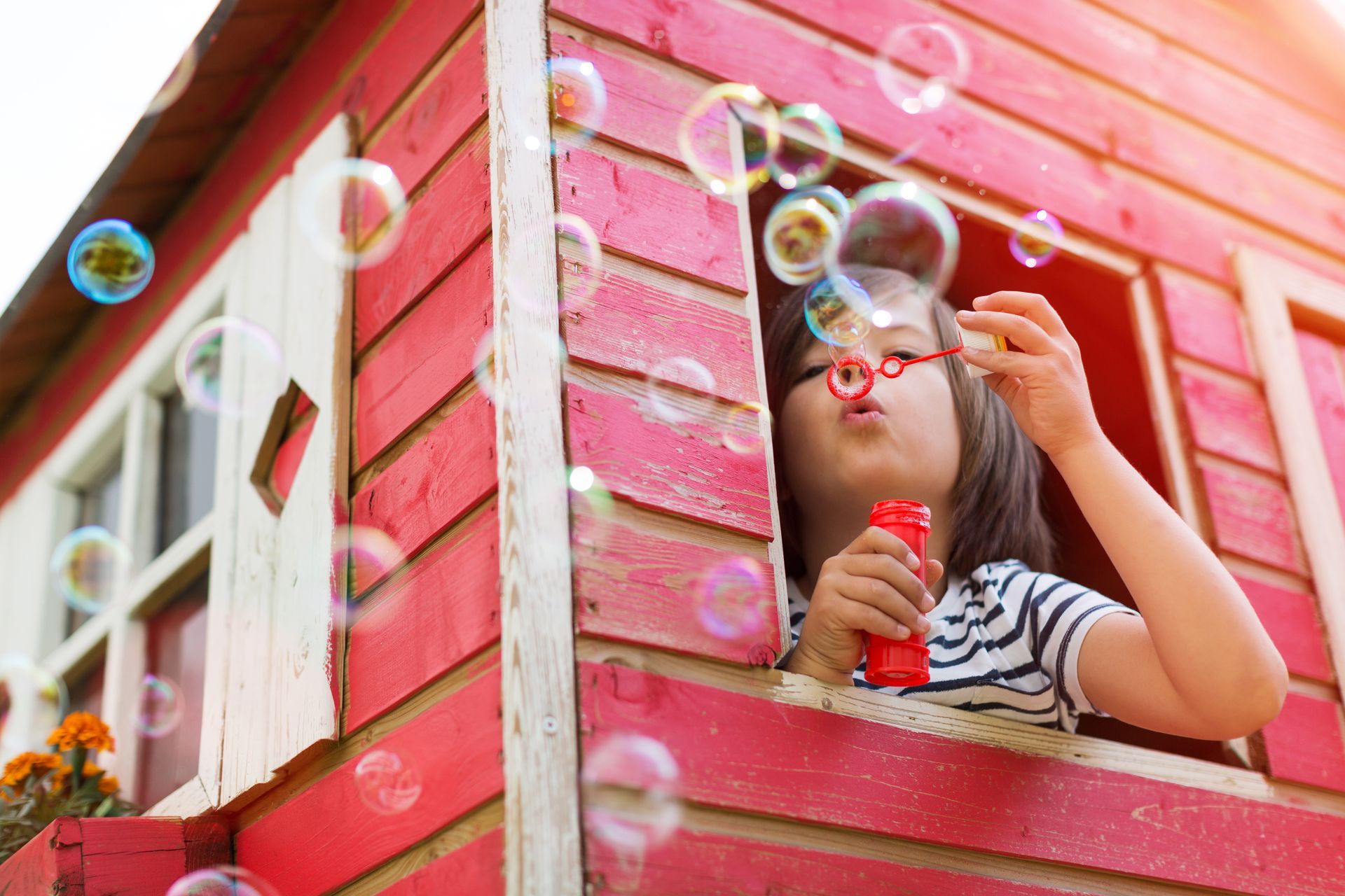 A little girl is blowing soap bubbles out of a window in a playhouse.