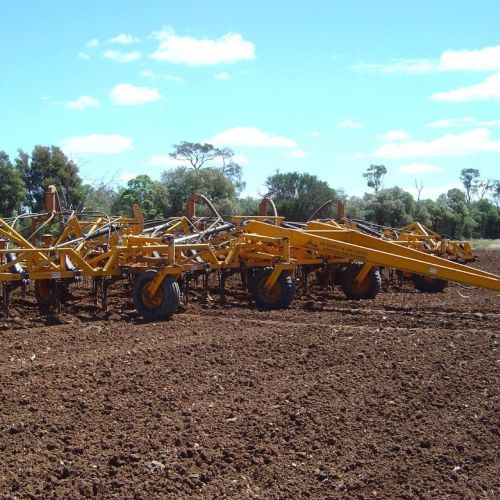 Yellow farming implement on brown soil under a blue sky with trees.