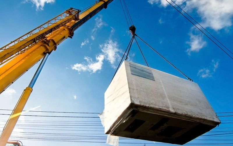 A yellow crane lifting a large, wrapped box against a blue sky with power lines.
