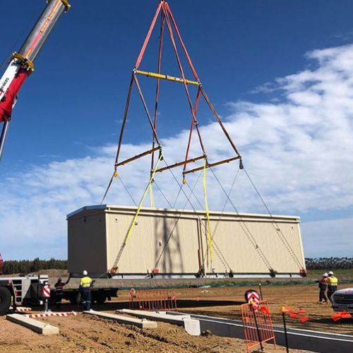 Crane lifting a tan building module from a truck bed at a construction site under a blue sky.