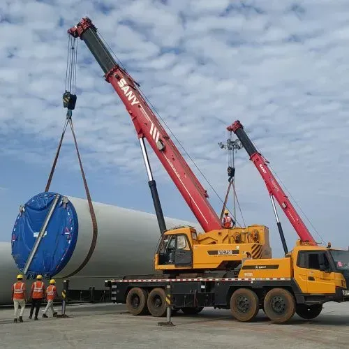 Two cranes lifting a wind turbine component. Workers in orange vests stand nearby.