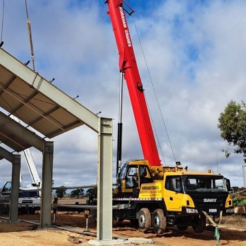 A red crane lifting a metal beam on a construction site under a partly cloudy sky.
