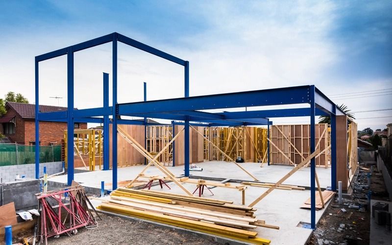Blue steel frame of a house under construction, with wood framing and materials on the ground.