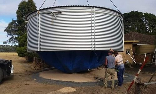 Large water tank being lowered into place by a crane; two men assist on the ground, rural setting.