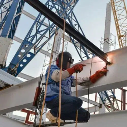 Welder in protective gear working on steel beams at a construction site, with blue and yellow structures in background.