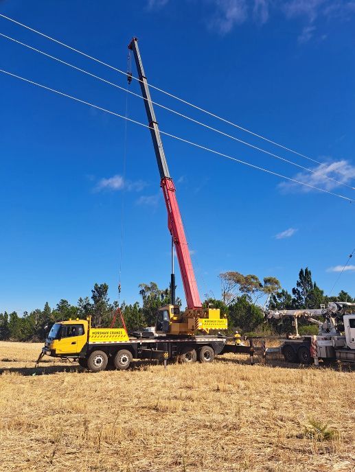 A crane positioned near power lines in a field on a sunny day. The boom is extended upward, reaching the lines.