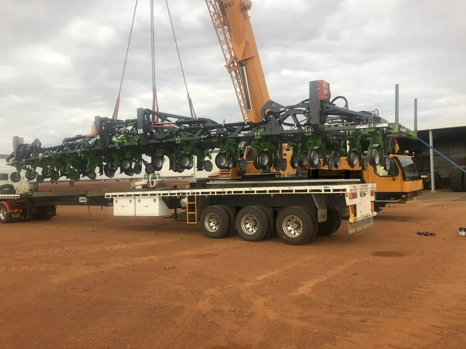 A crane lifting a large agricultural planter onto a flatbed trailer on a dirt surface.