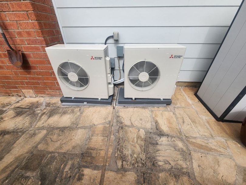 Two white outdoor air conditioner units beside a brick wall on a patio.