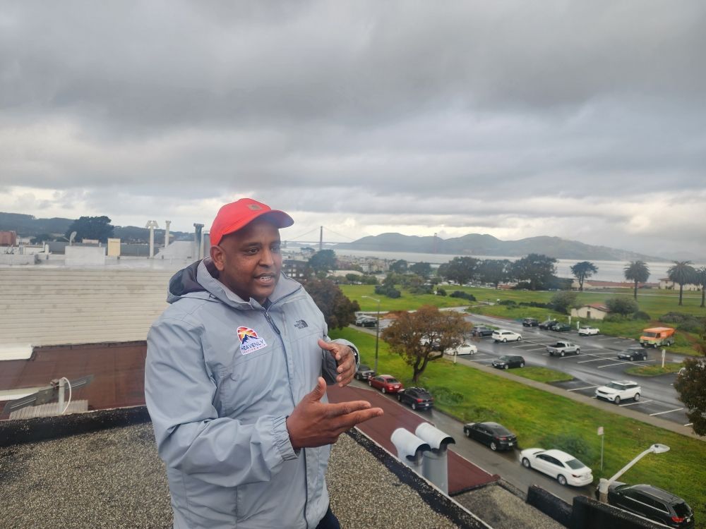 Man in a red cap and gray jacket standing on a rooftop, gesturing over a cloudy waterfront view
