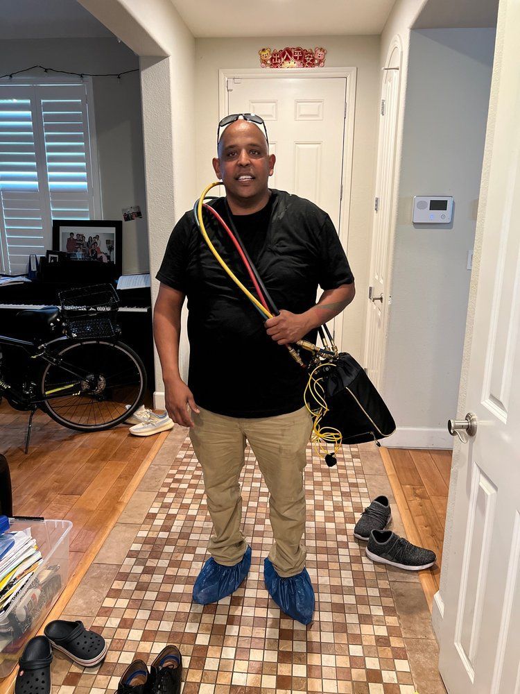 Person standing in a home entryway, wearing a black shirt, tan pants, blue shoe covers, and a crossbody bag.