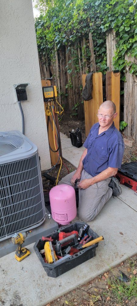 Person kneeling beside tools and a pink container, working outdoors near a fence and plants.