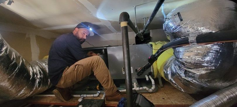 Technician inspecting ductwork in a crawl space with insulation and exposed pipes