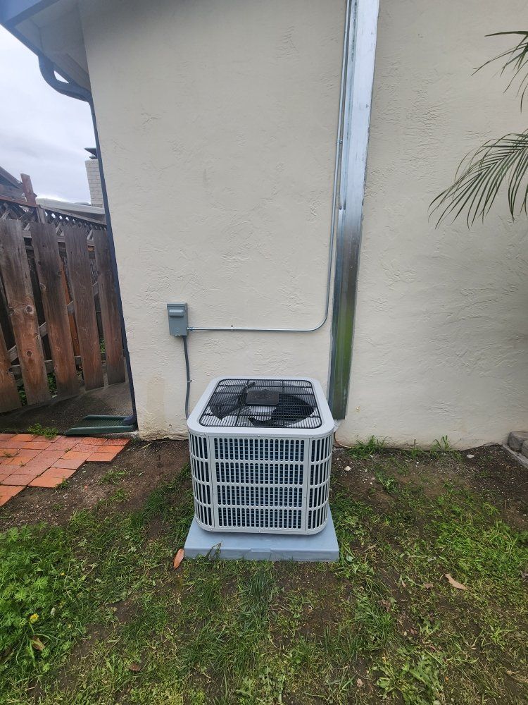 Outdoor air conditioner unit against a beige house wall with a wooden fence nearby.