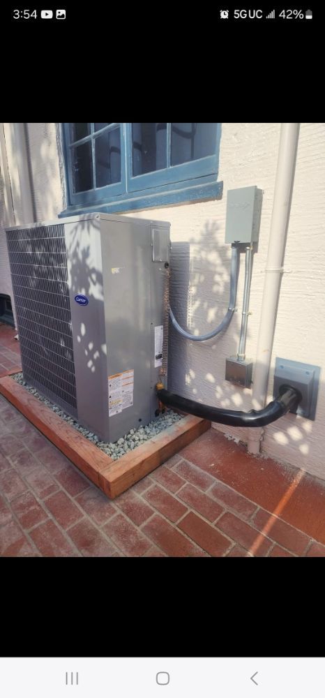 Outdoor air-conditioning unit beside a brick wall, with black piping and a small utility box.