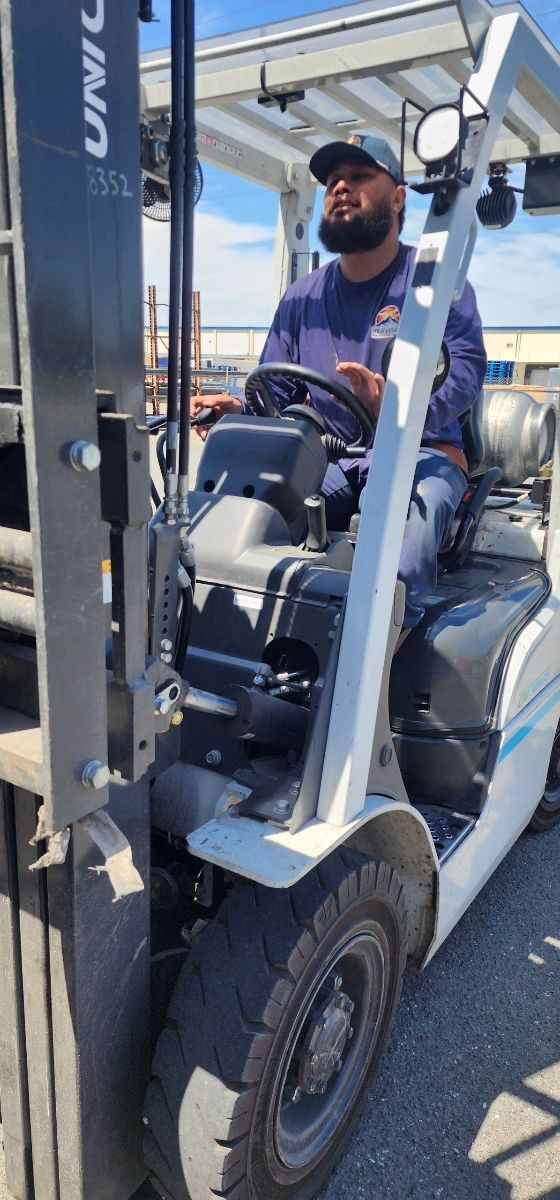 Man driving a blue forklift outdoors in a parking lot, wearing a navy cap and shirt.