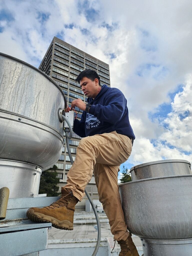 Worker in blue jacket and tan pants inspecting rooftop equipment beside a tall building.