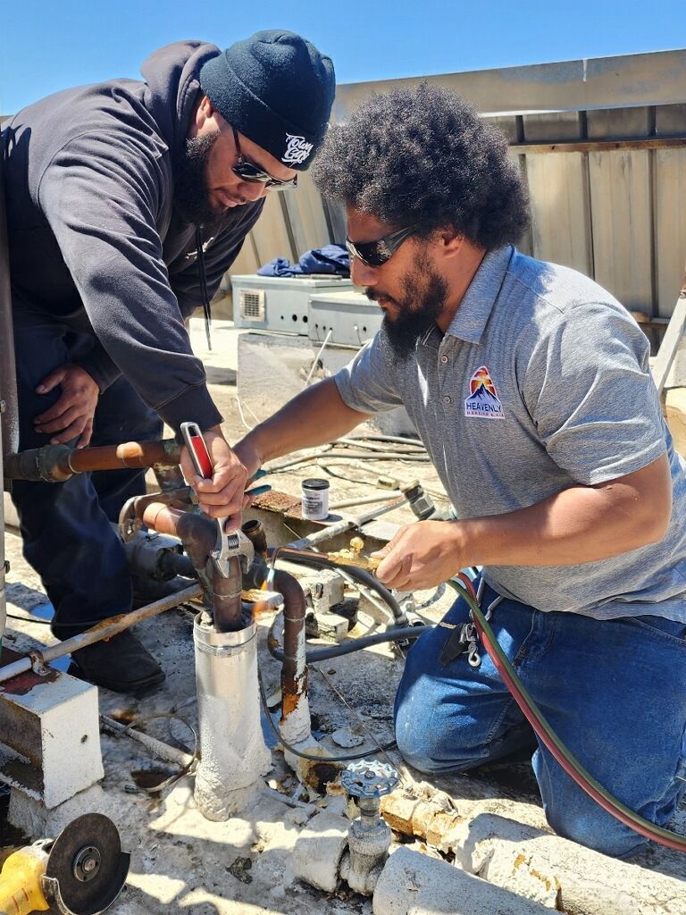 Two workers crouch over metal piping on a rooftop, using tools to repair equipment.